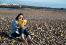 woman in blue denim jacket sitting on rocky shore reading book during daytime